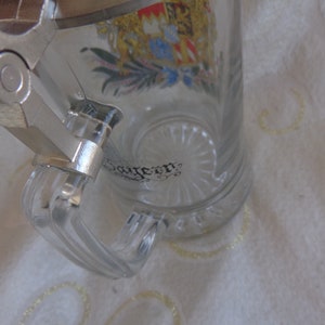 May include: A clear glass beer stein with a silver lid and handle. The stein has a colorful crest with a crown and a floral design. The text "Bayern" is visible on the stein.