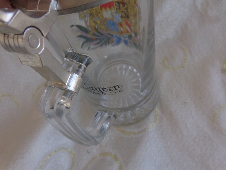 May include: A clear glass beer stein with a silver lid and handle. The stein has a colorful crest with a crown and a floral design. The text "Bayern" is visible on the stein.