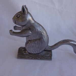 May include: A silver-toned metal nutcracker shaped like a squirrel sitting on a textured base. The squirrel is depicted in profile with its tail extended horizontally, featuring molded fur details and a functional lever mechanism for cracking nuts.