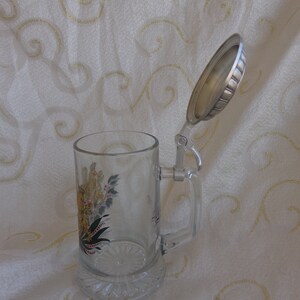 May include: A clear glass beer stein with a hinged lid. The stein has a colorful floral design and a silver-colored handle. The lid is open and is made of metal.