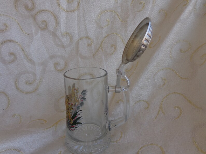 May include: A clear glass beer stein with a hinged lid. The stein has a colorful floral design and a silver-colored handle. The lid is open and is made of metal.