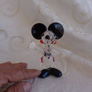 May include: A glass figurine of Mickey Mouse with black ears, red cheeks, and a black and white outfit. The figurine is standing on a white surface.