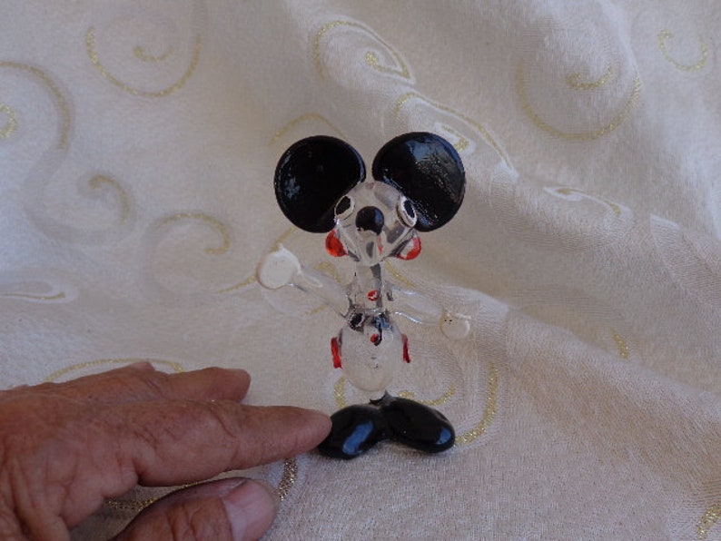 May include: A glass figurine of Mickey Mouse with black ears, red cheeks, and a black and white outfit. The figurine is standing on a white surface.