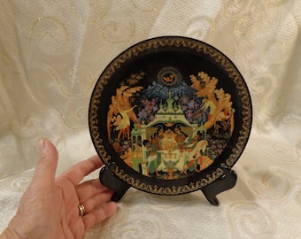 May include: A black and gold decorative plate with a colorful painted scene of people and trees. The plate is on a black stand.