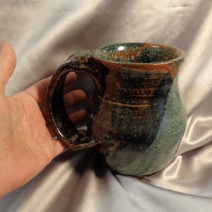 May include: Handmade ceramic mug with a dark brown handle and rim, and a blue and green body. The mug has a unique, organic shape and a glossy finish. A perfect addition to any kitchen.
