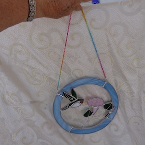 May include: A stained glass suncatcher with a blue background, featuring a hummingbird and a pink flower. The suncatcher is hanging from a rainbow-colored chain.