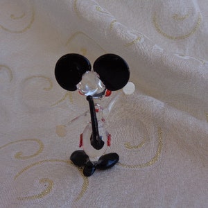 May include: A glass figurine of a mouse with black ears and red accents. The mouse is standing on a white surface.
