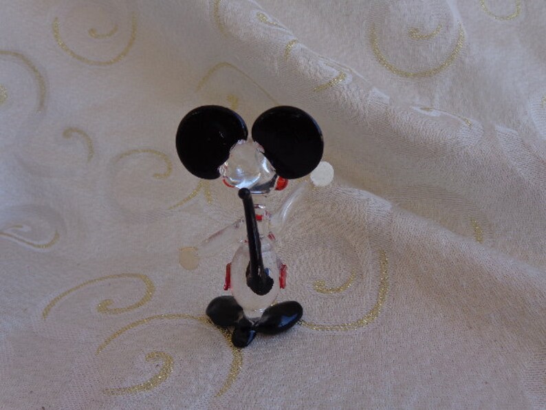 May include: A glass figurine of a mouse with black ears and red accents. The mouse is standing on a white surface.
