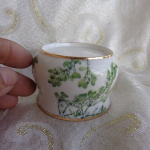 May include: A small, white ceramic container with a gold rim and base. The container is decorated with green floral designs. The rim and base have a gold trim. The container is held by a hand.