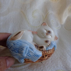 May include: A white ceramic figurine of a cat sleeping in a blue and white striped basket. The cat has pink ears and a white face with black eyes.