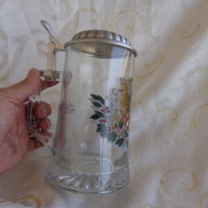 May include: A glass beer stein with a pewter lid and a handle. The stein has a painted design of a winter scene with holly and berries.