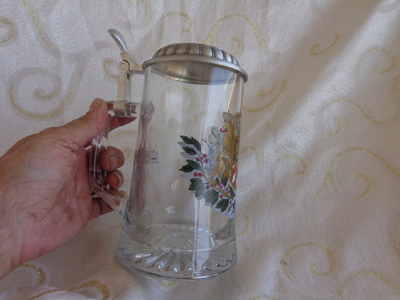May include: A glass beer stein with a pewter lid and a handle. The stein has a painted design of a winter scene with holly and berries.
