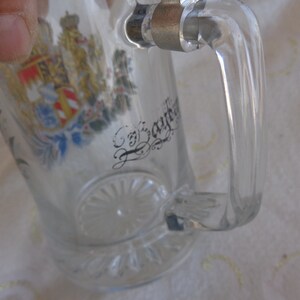 May include: Clear glass beer stein with a handle. The stein has a colorful coat of arms and the text "Bayern" on the front.
