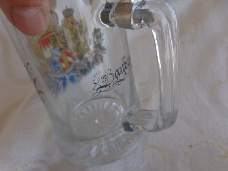 May include: Clear glass beer stein with a handle. The stein has a colorful coat of arms and the text "Bayern" on the front.