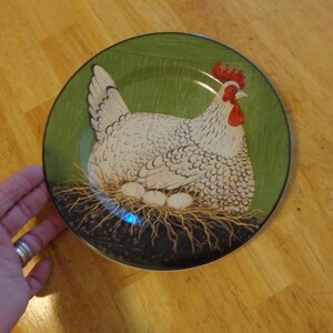 May include: A round decorative plate featuring a white hen with a red comb and wattle, sitting on a nest with three eggs. The background is green, and the plate has a black rim. The plate is on a wooden surface.