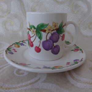 May include: A white ceramic teacup and saucer with a floral design featuring purple plums, red cherries, and yellow flowers. The cup has a handle and the saucer has a scalloped edge.