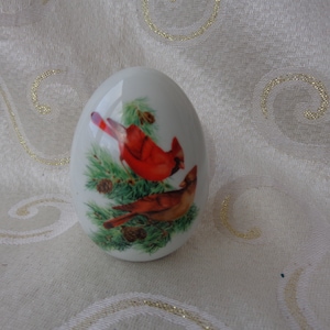 May include: White ceramic egg-shaped ornament with a painted image of two red cardinals perched on a pine branch with green needles and pine cones.