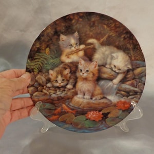 May include: A decorative ceramic plate featuring a whimsical scene of four kittens playing by a waterfall. The kittens are painted in shades of white, brown, and orange, and the background is a lush forest setting with green ferns and rocks.
