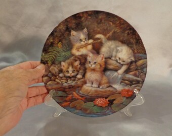 May include: A decorative ceramic plate featuring a whimsical scene of four kittens playing by a waterfall. The kittens are painted in shades of white, brown, and orange, and the background is a lush forest setting with green ferns and rocks.