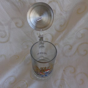 May include: A glass beer stein with a pewter lid. The stein features a colorful coat of arms with a crown and a lion. The lid has a small sticker that says "0.5L."