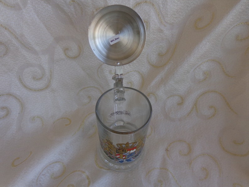 May include: A glass beer stein with a pewter lid. The stein features a colorful coat of arms with a crown and a lion. The lid has a small sticker that says "0.5L."