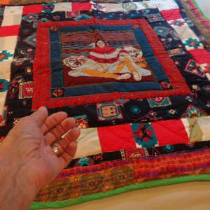 May include: A colorful handmade quilt with a central image of a person in a blanket. The quilt features red, black, and white geometric patterns. The border is green. The quilt is likely made of cotton or a similar fabric.