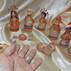 May include: A collection of hand-painted ceramic figurines, including angels, figures with headdresses, and animals. The figurines are in various colors, with detailed patterns and designs. The collection is arranged on a gold satin fabric.