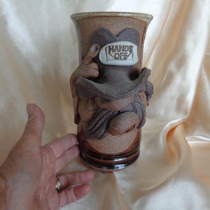 May include: A cylindrical brown ceramic mug featuring a sculpted figure wearing a wide-brimmed hat. The hat displays a white rectangular plaque with the embossed text HANDS OFF. The mug has a textured, speckled finish with a darker brown base.