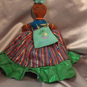 May include: A handmade doll with brown skin, black eyes, and a colorful striped skirt. The doll is wearing a blue shirt and a green bag with a palm tree design and the word "Jamaica" embroidered on it.