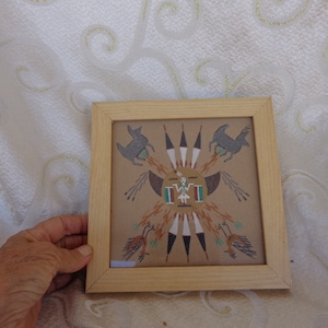 May include: A framed sand painting with a Native American design. The painting features a central figure with feathers and two animals on either side. The frame is made of light wood.