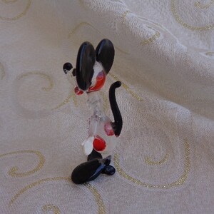 May include: A glass figurine of a cartoon mouse with black ears and a tail. The mouse is standing on its hind legs and has red spots on its body.