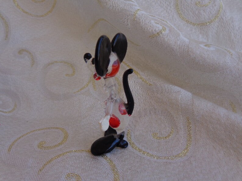 May include: A glass figurine of a cartoon mouse with black ears and a tail. The mouse is standing on its hind legs and has red spots on its body.