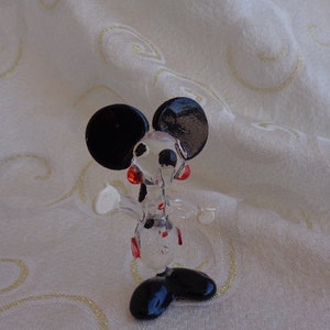 May include: A clear glass figurine of Mickey Mouse. The figurine is in a standing pose with black ears, black shoes, and red accents on the face.