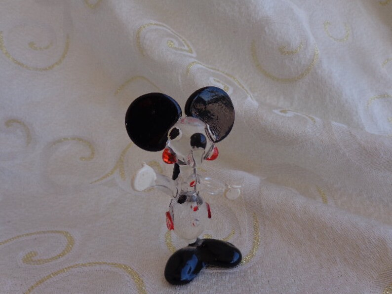 May include: A clear glass figurine of Mickey Mouse. The figurine is in a standing pose with black ears, black shoes, and red accents on the face.