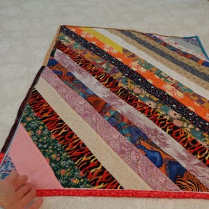 May include: A colorful patchwork quilt with a diagonal pattern of fabric strips in various colors and designs. The quilt is edged with a red border.