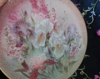 May include: A decorative plate featuring a floral design with irises and other flowers in shades of white, purple, pink, and gold. A butterfly is also depicted. The plate has a gold rim.