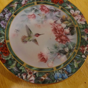 May include: Decorative plate featuring a hummingbird in flight, surrounded by pink and red flowers. The plate has a green border with additional hummingbird and floral designs. The rim is trimmed with gold. The plate is signed "LENA".