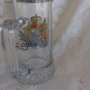 May include: A clear glass beer stein with a silver lid and a gold and colorful coat of arms with the word "Bayern" printed on it.