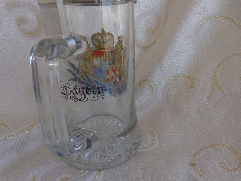 May include: A clear glass beer stein with a silver lid and a gold and colorful coat of arms with the word "Bayern" printed on it.