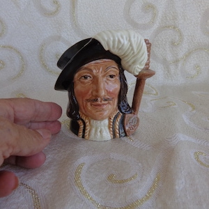 May include: A ceramic figurine of a man wearing a black hat with a white feather plume. He has a brown beard and mustache and is wearing a brown and white patterned jacket. The figurine is holding a staff with a white feather plume.