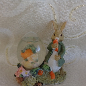 May include: A ceramic figurine of a rabbit holding a carrot, standing next to a clear glass dome with a floral design and a small carrot inside. The figurine and dome are on a base with green leaves and flowers.