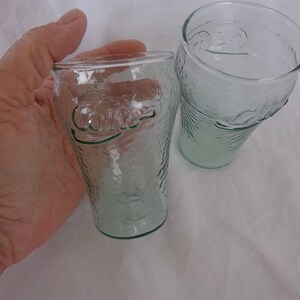 May include: Two clear glass Coca-Cola glasses with the Coca-Cola logo embossed on the side. The glasses have a textured surface.