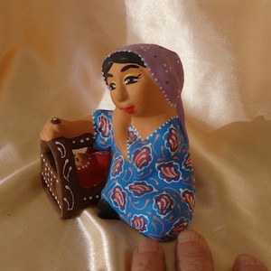May include: A hand-painted ceramic figurine of a woman wearing a blue dress with floral patterns and a purple head covering. The figurine is holding a small brown cradle. The background is a satin fabric.
