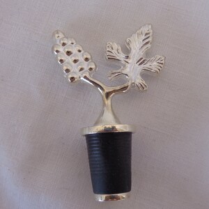 May include: Silver wine bottle stopper with a grapevine design. The stopper has a black rubber base.