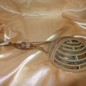 May include: A ceramic pendant featuring a rounded, dome-like shape with a raised, labyrinthine pattern in shades of green, blue, and brown. The pendant is attached to a brown cord adorned with small, light-colored, irregularly shaped beads.