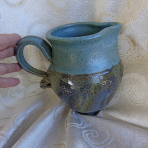 May include: A handmade ceramic pitcher with a blue-green glaze and brown accents. The pitcher has a wide mouth and a handle.
