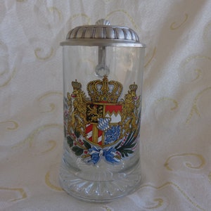 May include: A clear glass beer stein with a silver lid and a colorful coat of arms featuring a crown, lions, and a shield with red and white stripes.