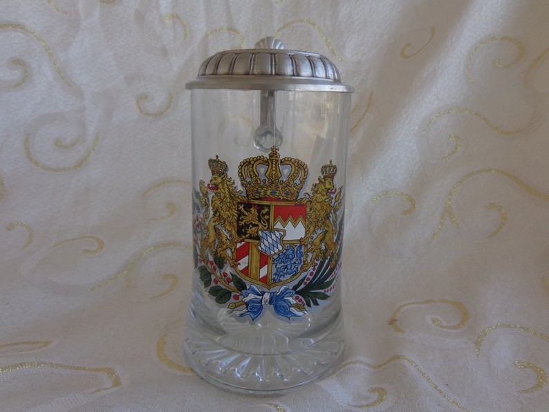 May include: A clear glass beer stein with a silver lid and a colorful coat of arms featuring a crown, lions, and a shield with red and white stripes.