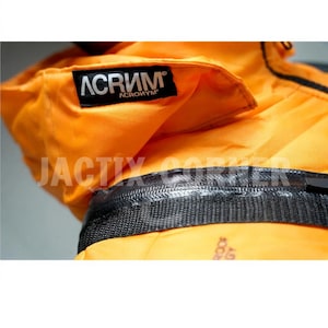 May include: Close-up of a bright orange jacket with a black zipper and a black label that says "ACRONYM". The jacket has a textured, woven appearance and a folded collar. The text "JACTIX CORNER" is visible.