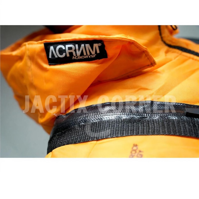 May include: Close-up of a bright orange jacket with a black zipper and a black label that says "ACRONYM". The jacket has a textured, woven appearance and a folded collar. The text "JACTIX CORNER" is visible.